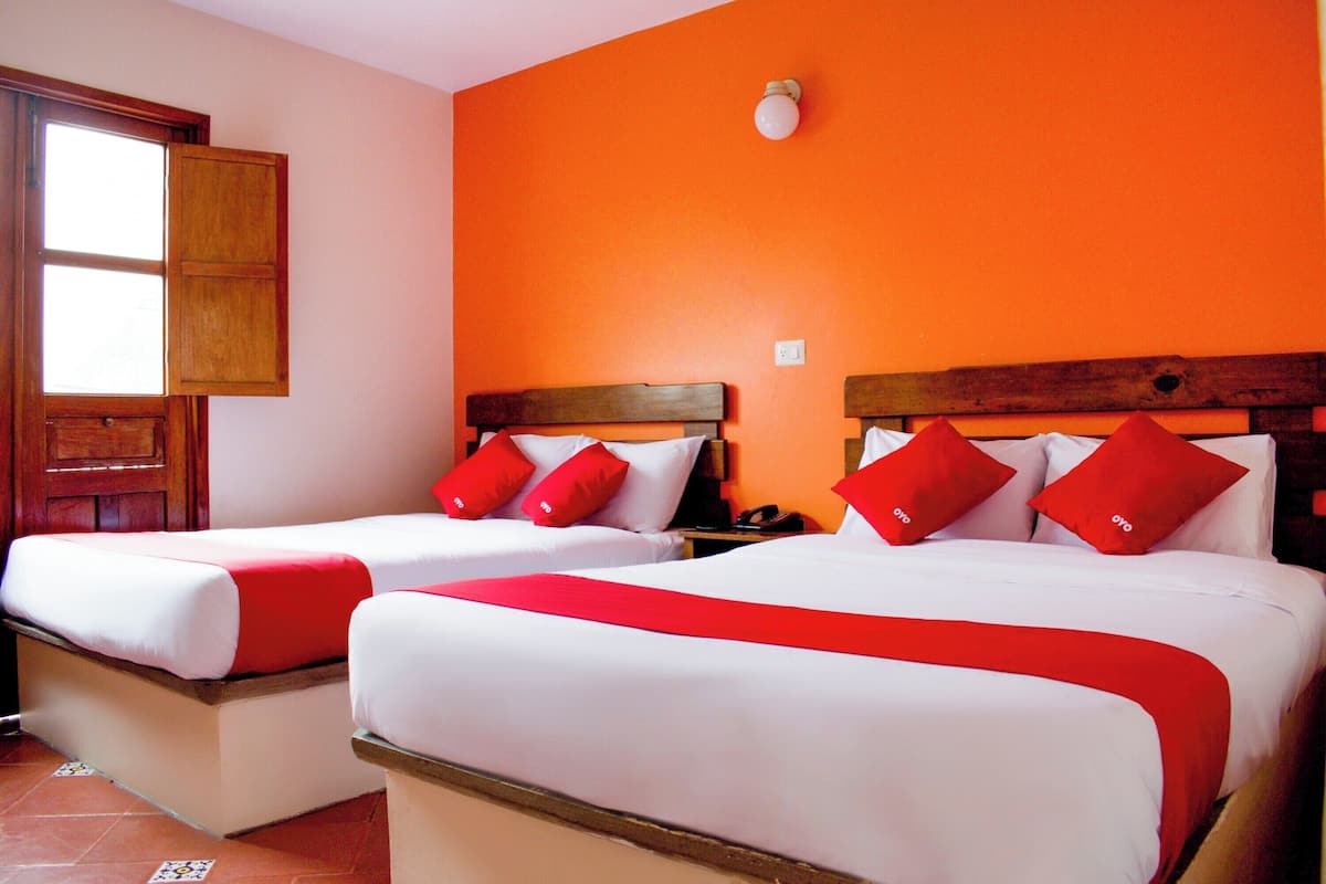 Dos camas con ropa de cama blanca y roja, un cabecero de madera, una ventana con marco de madera y una pared anaranjada.