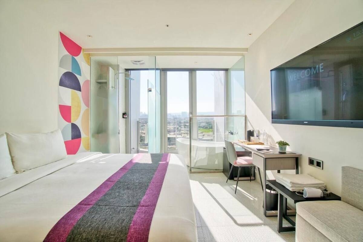 Una habitación de hotel moderna con una cama grande, un escritorio, una silla, un televisor y un balcón con vista a la ciudad.