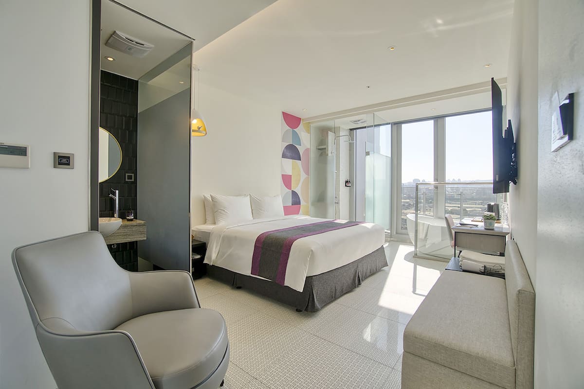 Una habitación de hotel moderna con cama, escritorio, silla y un amplio ventanal con vistas a la ciudad.