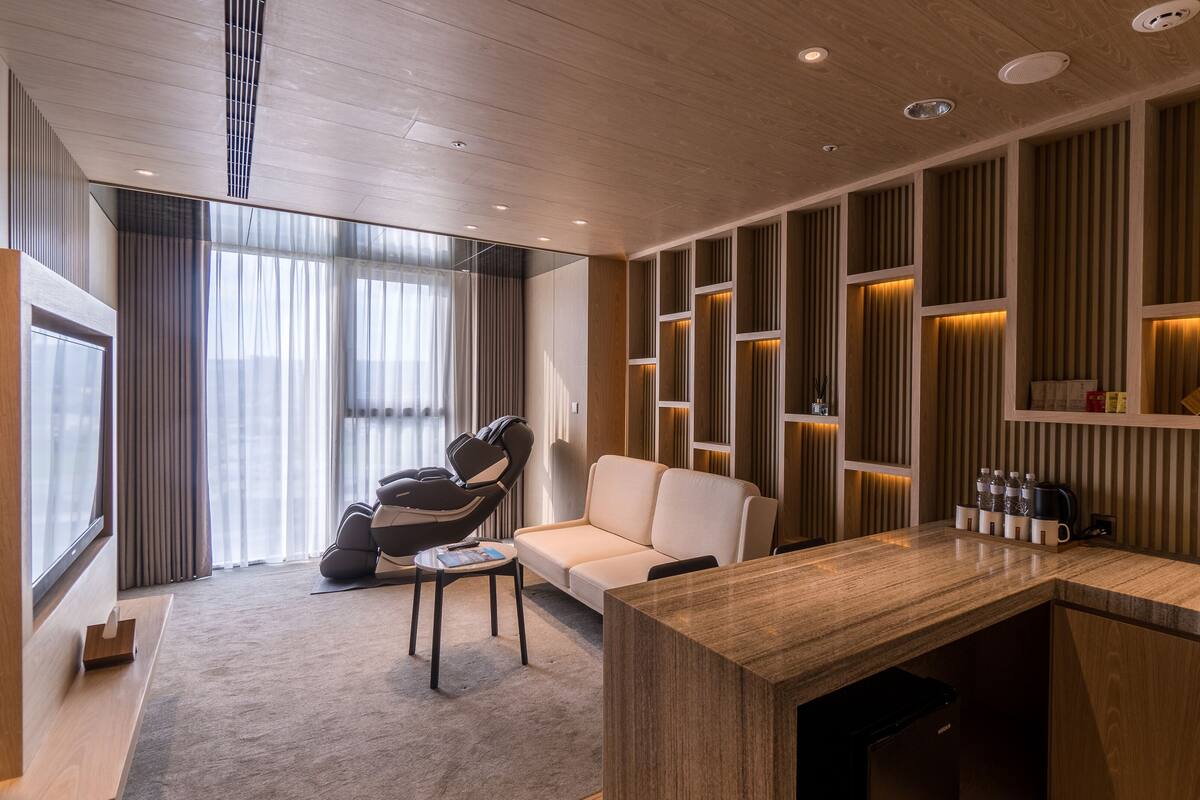 Una habitación de hotel moderna con sillón de masajes, sofá, una mesita y una zona de cocina con fregadero y encimera.
