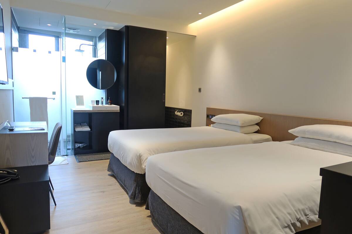 Habitación de hotel con dos camas, un escritorio, una silla, un espejo y un baño con ducha.