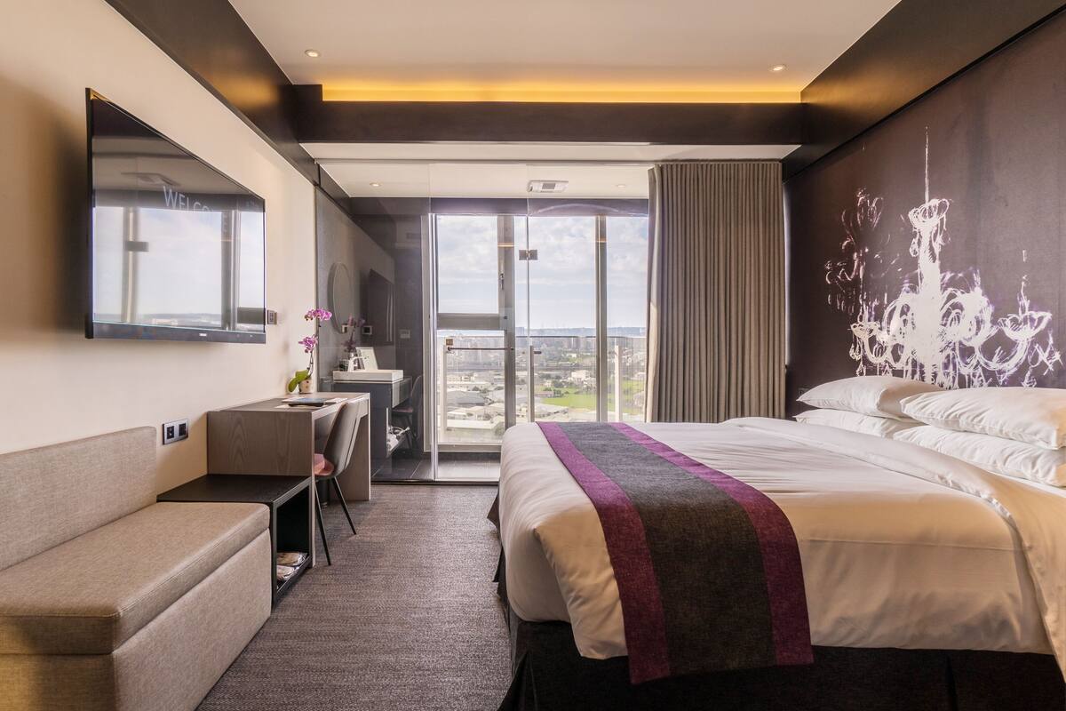 Habitación de hotel con una cama grande, un escritorio, una silla y vista de la ciudad a través de un ventanal.