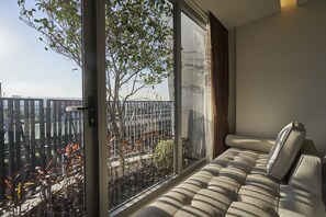 A modern living room with a large window, a sofa, and a view of a balcony with plants and a fence.
