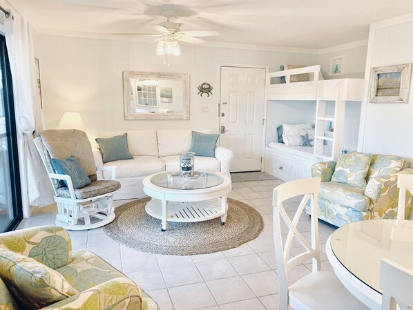 A well-lit living room with a ceiling fan, white furniture, a round coffee table, and a large mirror on the wall.