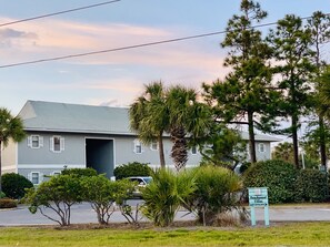 A two-story building with a grey roof and white trim, surrounded by palm trees and other greenery.