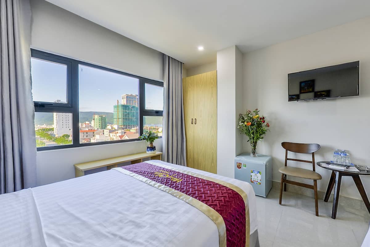 A hotel room with a large bed, a desk, a chair, a TV, and a window with a city view.