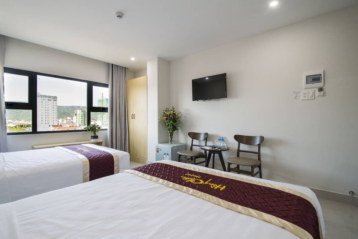A hotel room with two beds, a TV, a desk with chairs, and a window with a city view.