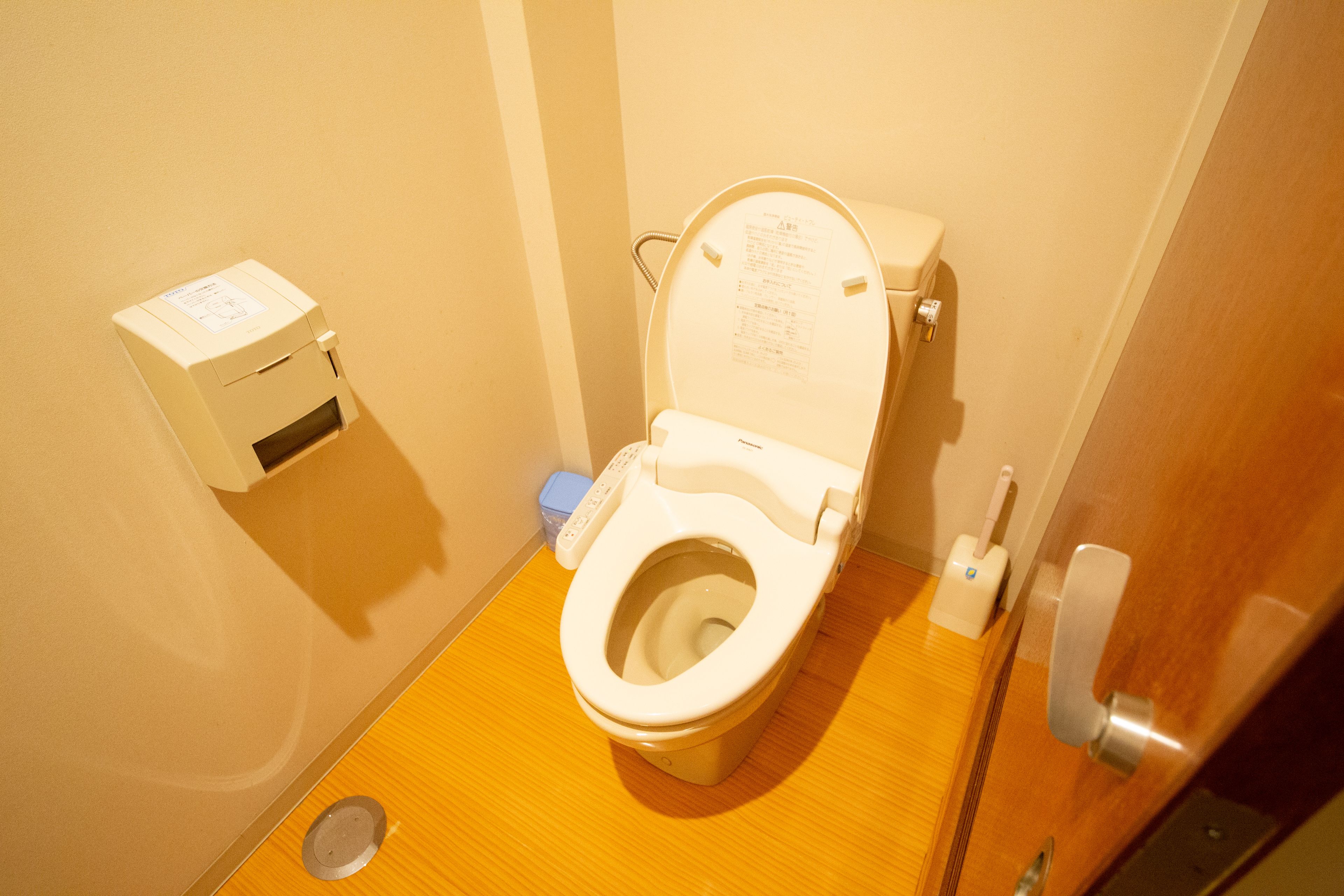 Un baño de hotel estándar con inodoro, dispensador de toallas de papel y un cubo de basura.