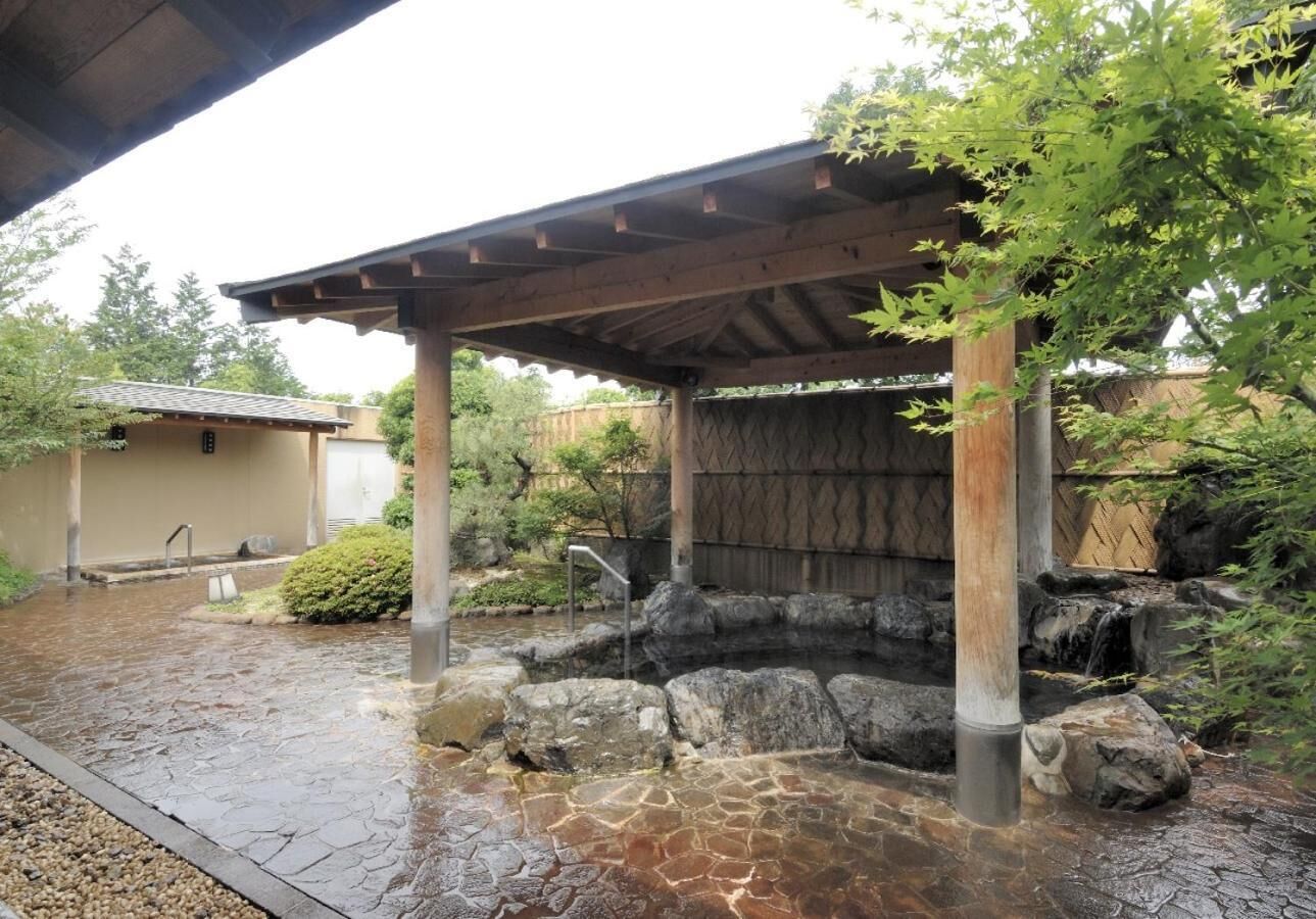 A traditional Japanese garden with a stone water feature, a wooden pavilion, and a small building in the background.