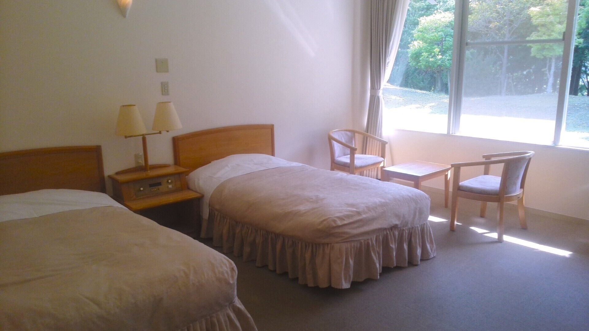 Ein Hotelzimmer mit zwei Betten, einem Schreibtisch, einem Stuhl und einem Fenster mit Blick auf Bäume.