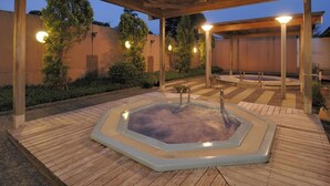 Una vasca idromassaggio sotto un pergolato di legno con un pavimento piastrellato, immersa nel verde e con un edificio sullo sfondo.