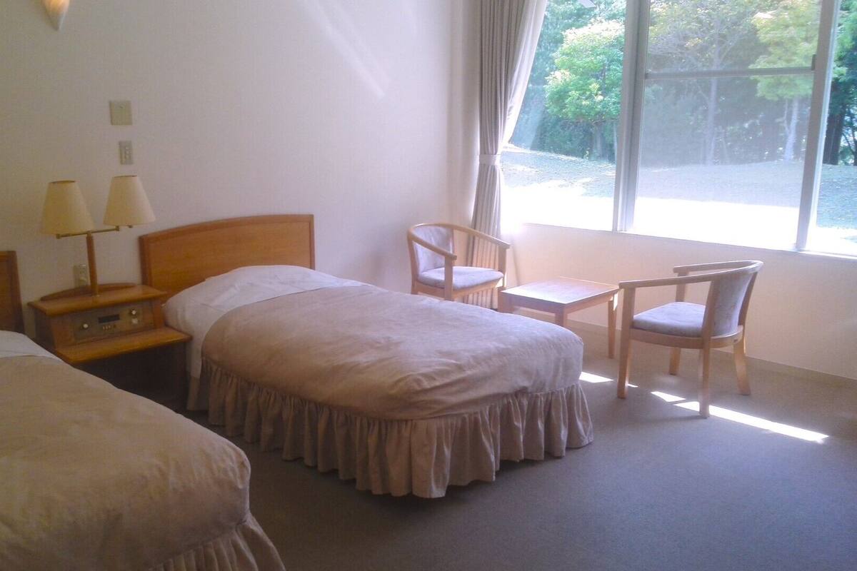 A hotel room with two single beds, a small table, a chair, and a window with a view of trees.