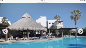 Een zwembadterras van een resort met een rieten overkapping, ligbedden en palmbomen.