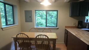 A dining area with a wooden table and chairs, two windows with a view of trees, and a kitchen with wooden cabinets and a countertop. - Quaint cabin with Lake Pauline views (Ludlow)