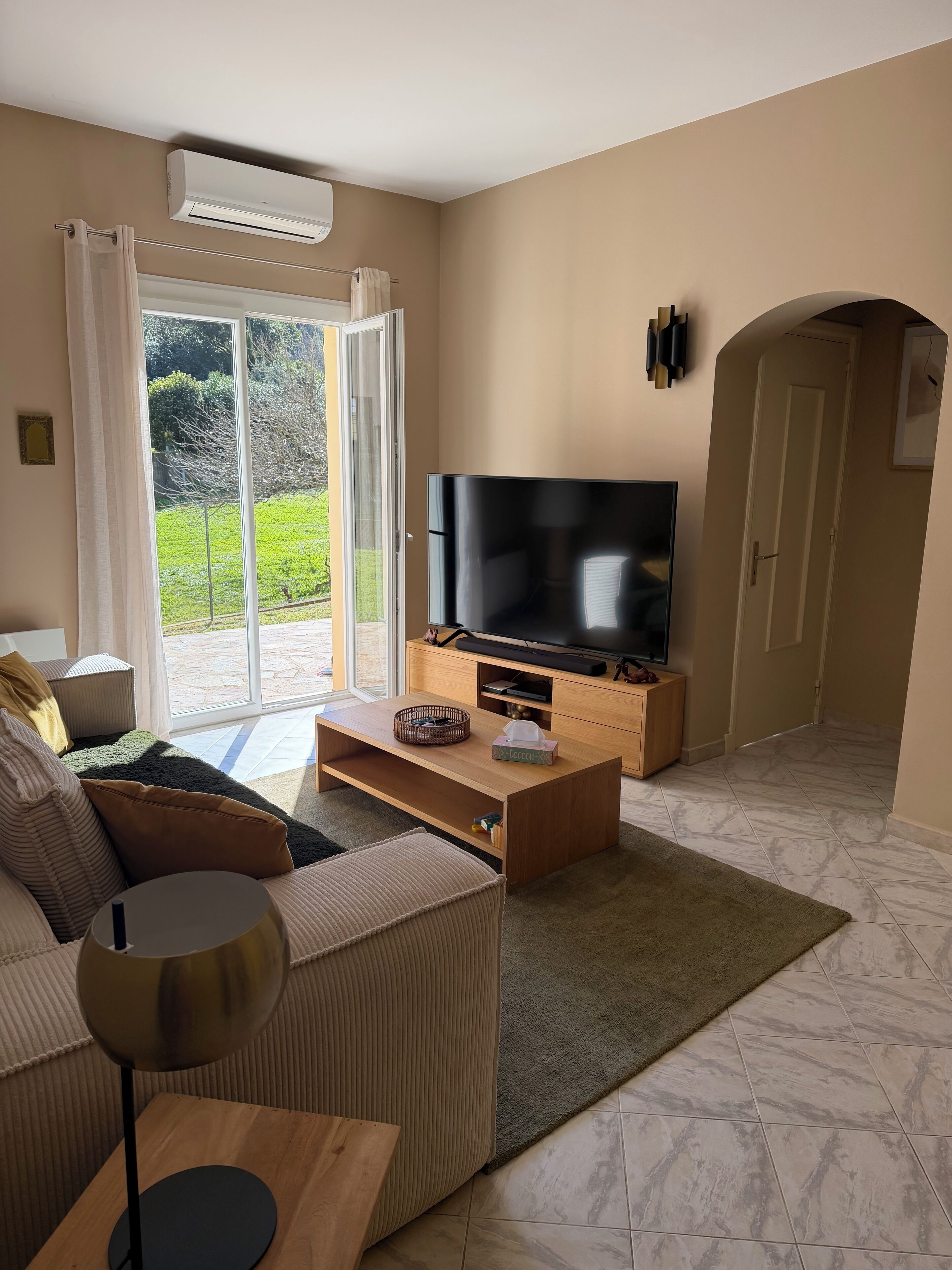 A living room with a sofa, a coffee table, a flat-screen TV, and a sliding glass door leading to a garden.