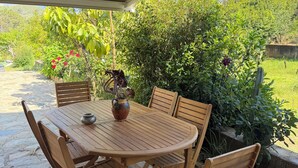 A wooden table and chairs under a covered patio, surrounded by greenery and a stone floor.