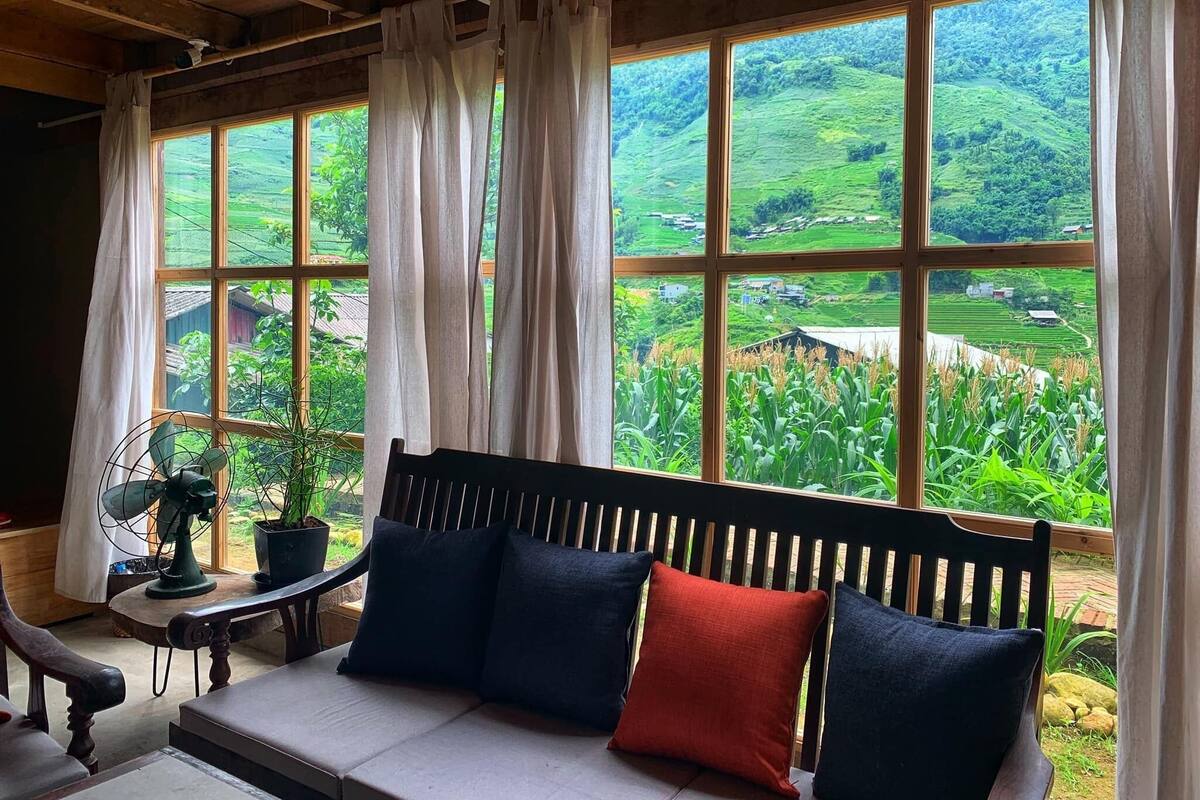 A room with a wooden bench, a table with a fan, and a view of green fields and houses through large windows.