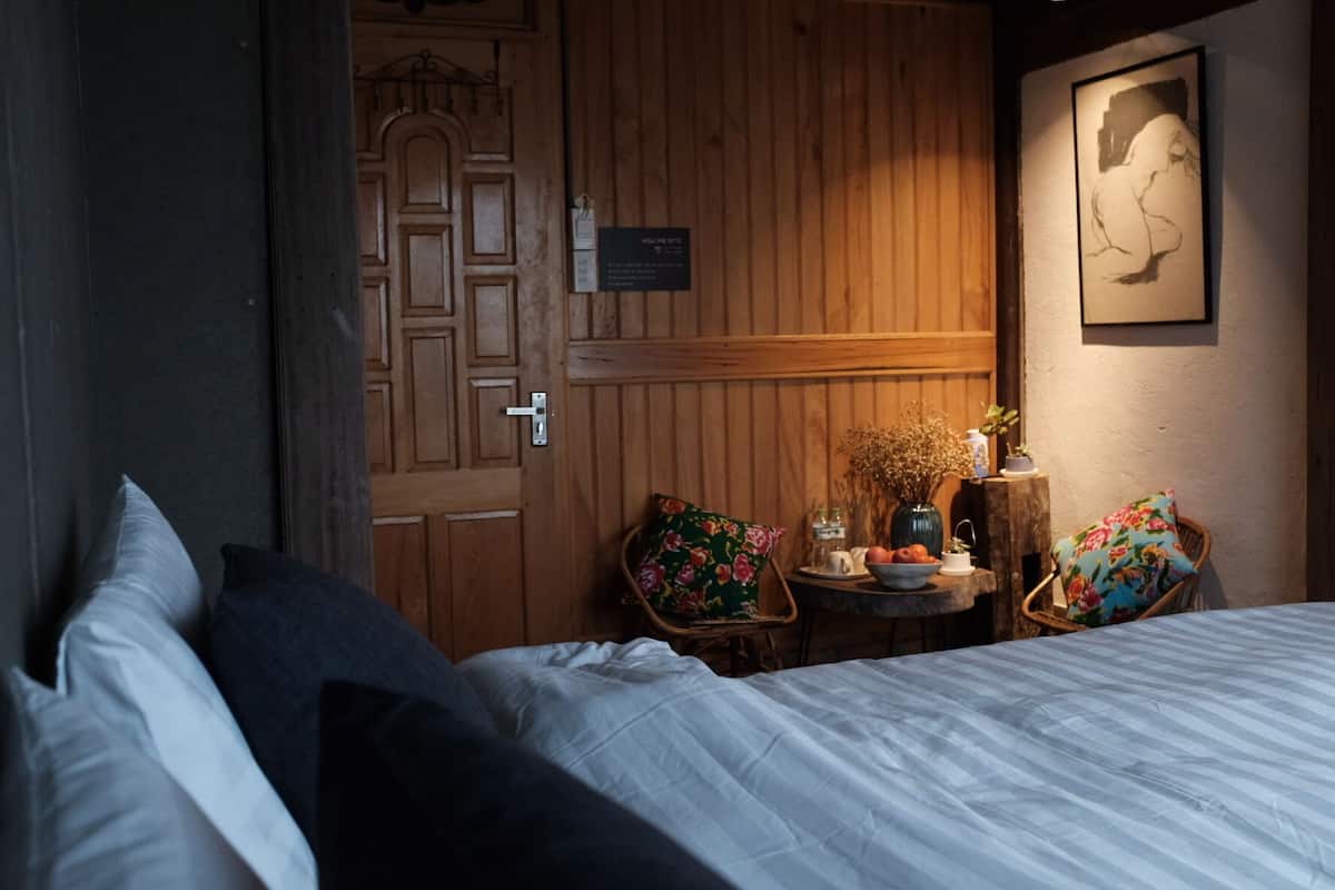 A hotel room with a bed, a wooden door, a small table with flowers, and a chair with a floral cushion.