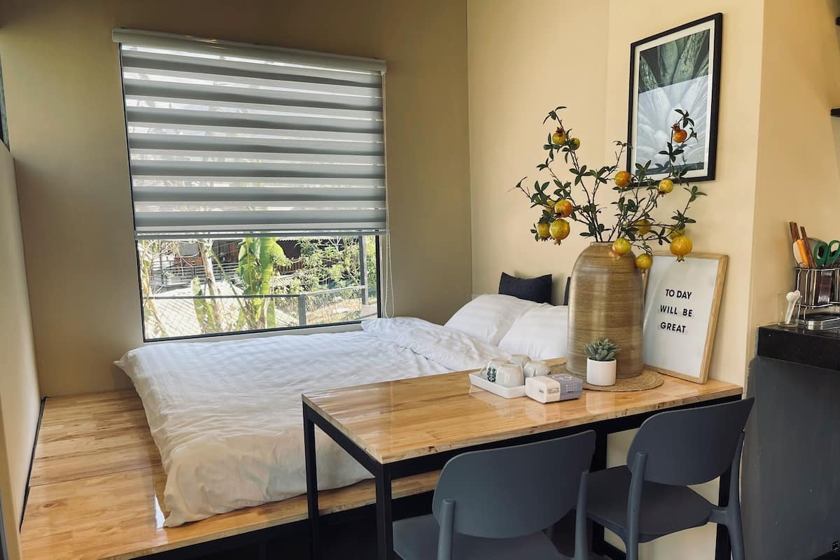 A small, cozy room with a bed, a wooden table, two chairs, a window with blinds, a potted plant, and a motivational quote on the wall.