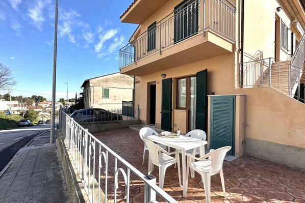 Un immeuble avec un balcon, une table entourée de chaises et une voiture garée à proximité.