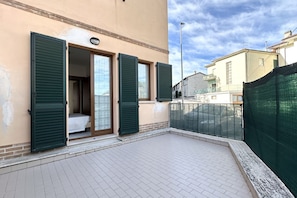 A building with a tiled patio, a balcony with green shutters, and a view of neighboring houses.