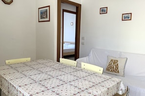 A room with a bed, a table with chairs, a door leading to another room, and a wall with framed pictures.