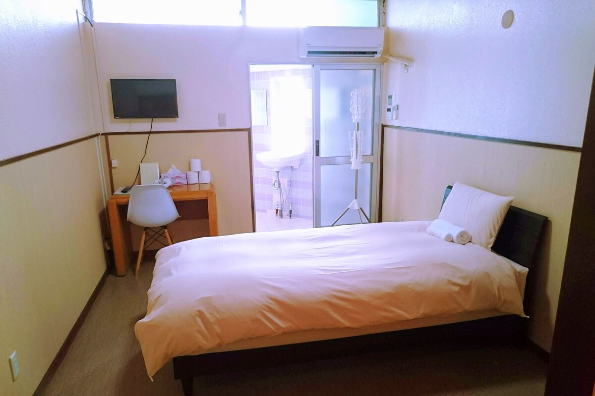 A hotel room with a bed, a desk, a chair, a TV, and a bathroom visible through an open door.