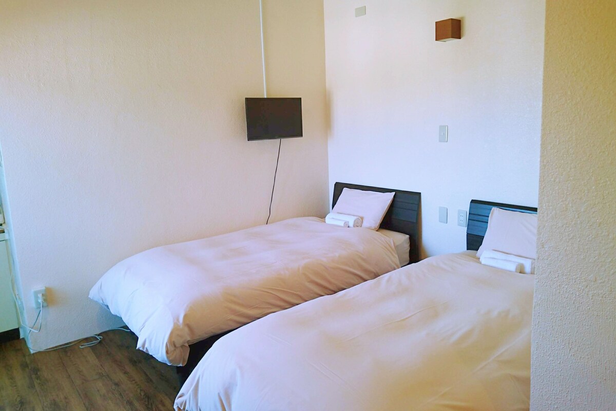 A simple, clean room with two single beds, a flat-screen TV mounted on the wall, and a small wooden shelf.