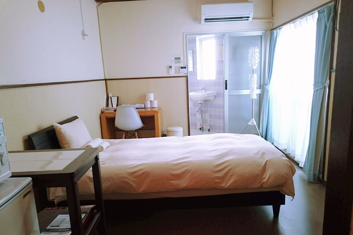 A hotel room with a bed, a desk, a chair, a small table, a window with curtains, and a bathroom visible through an open door.
