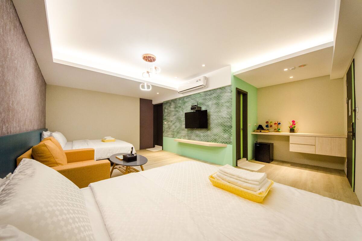 A modern hotel room with a bed, a sofa, a small table, and a kitchenette.