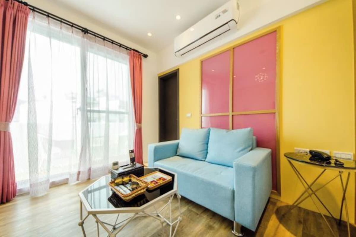 Una habitación con un sofá azul claro, una mesa de centro de vidrio, una mesita con un teléfono y una lámpara, y una ventana con cortinas rosadas.