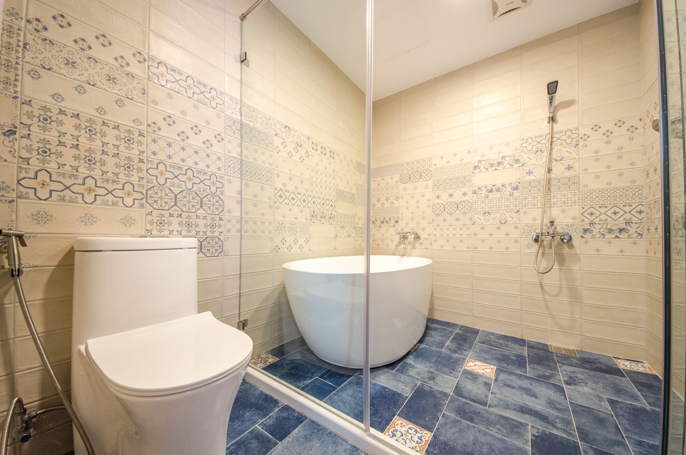 Et badeværelse med toilet, badekar og bruser. Væggene er beklædt med mønstrede fliser, og gulvet har blå fliser.