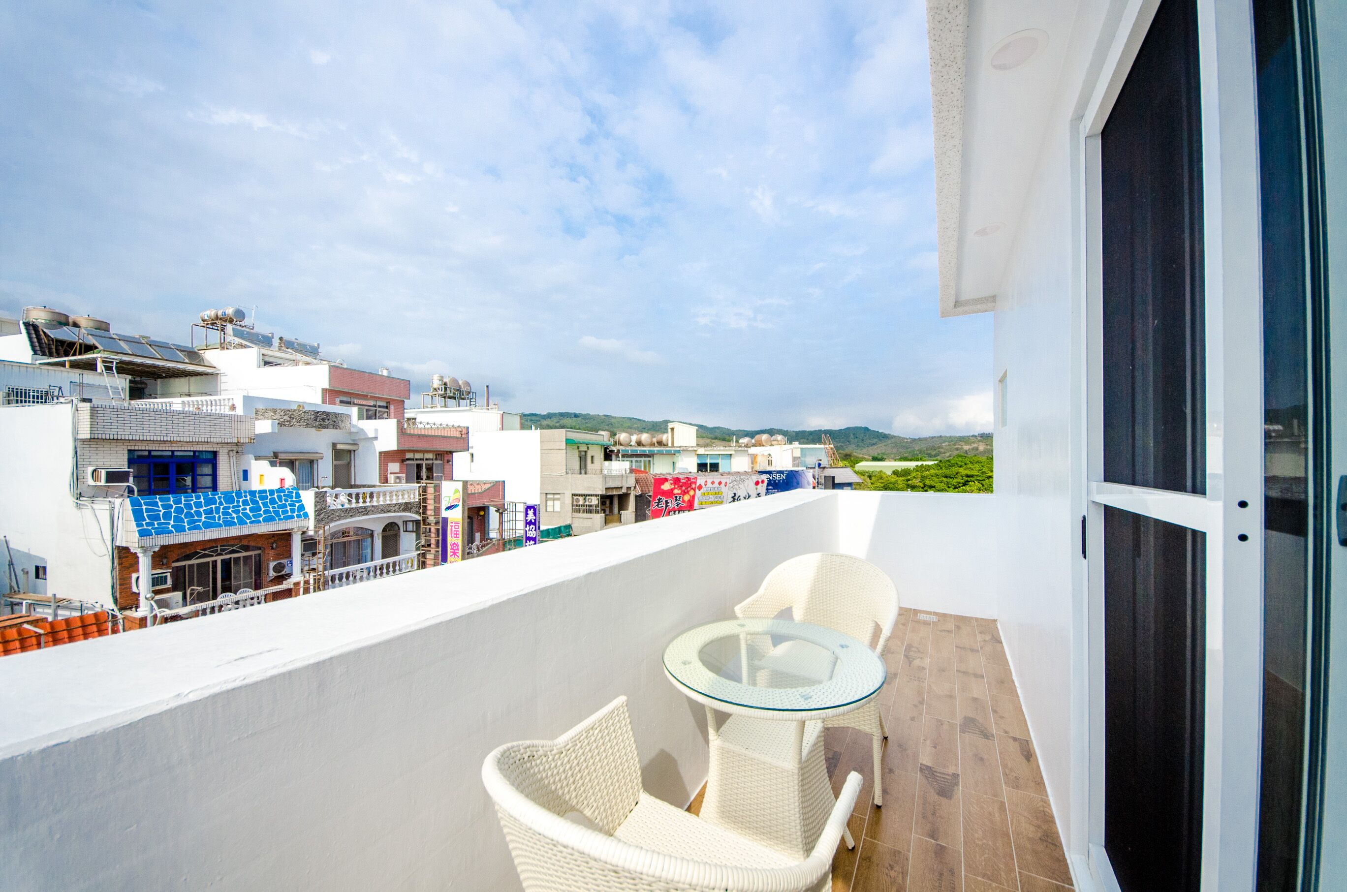 Un balcon avec une table en verre et des chaises en osier, offrant une vue sur un paysage urbain avec des bâtiments et de la verdure.