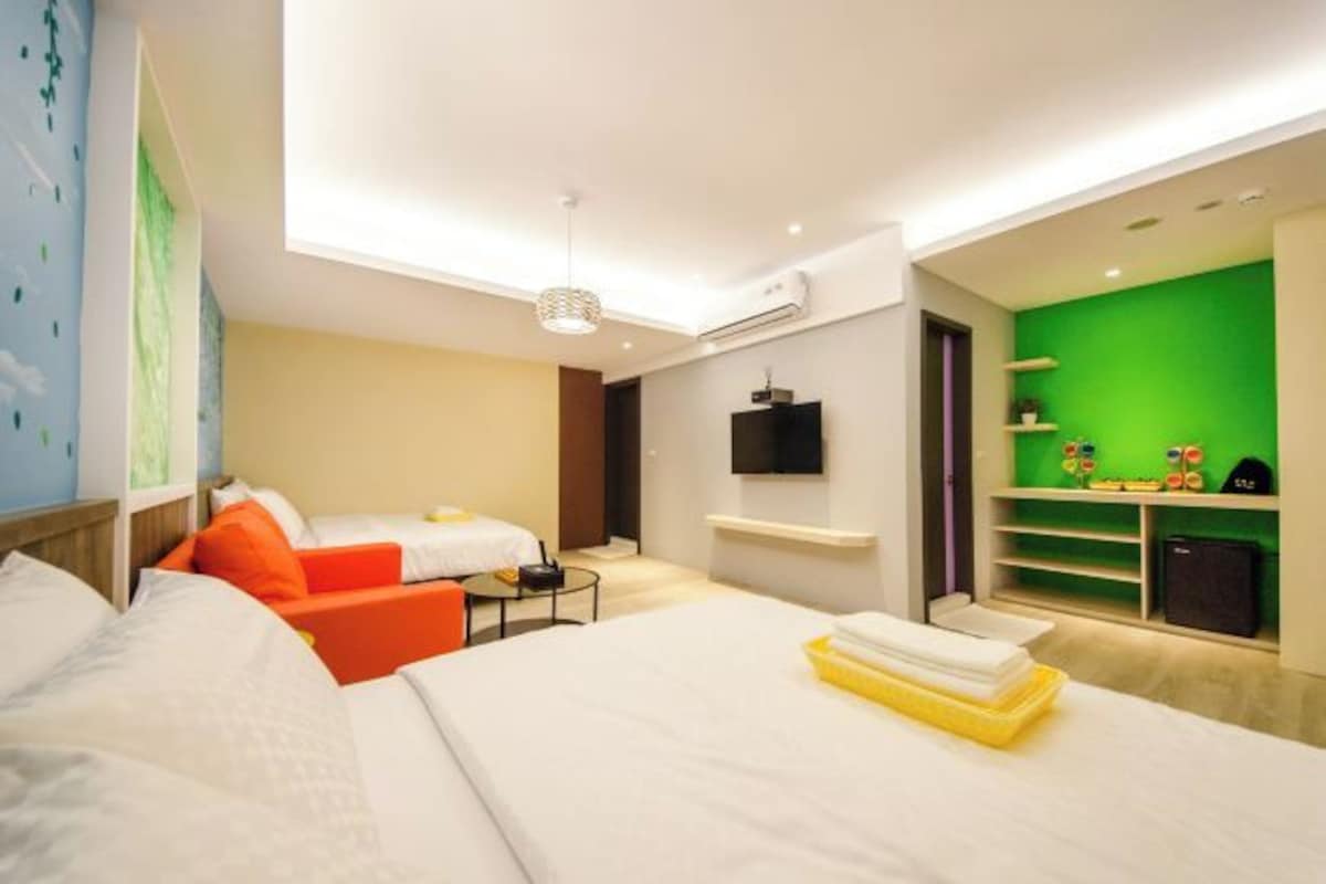 Habitación de hotel con cama, sofá, una mesita, televisor y pared verde con estanterías.