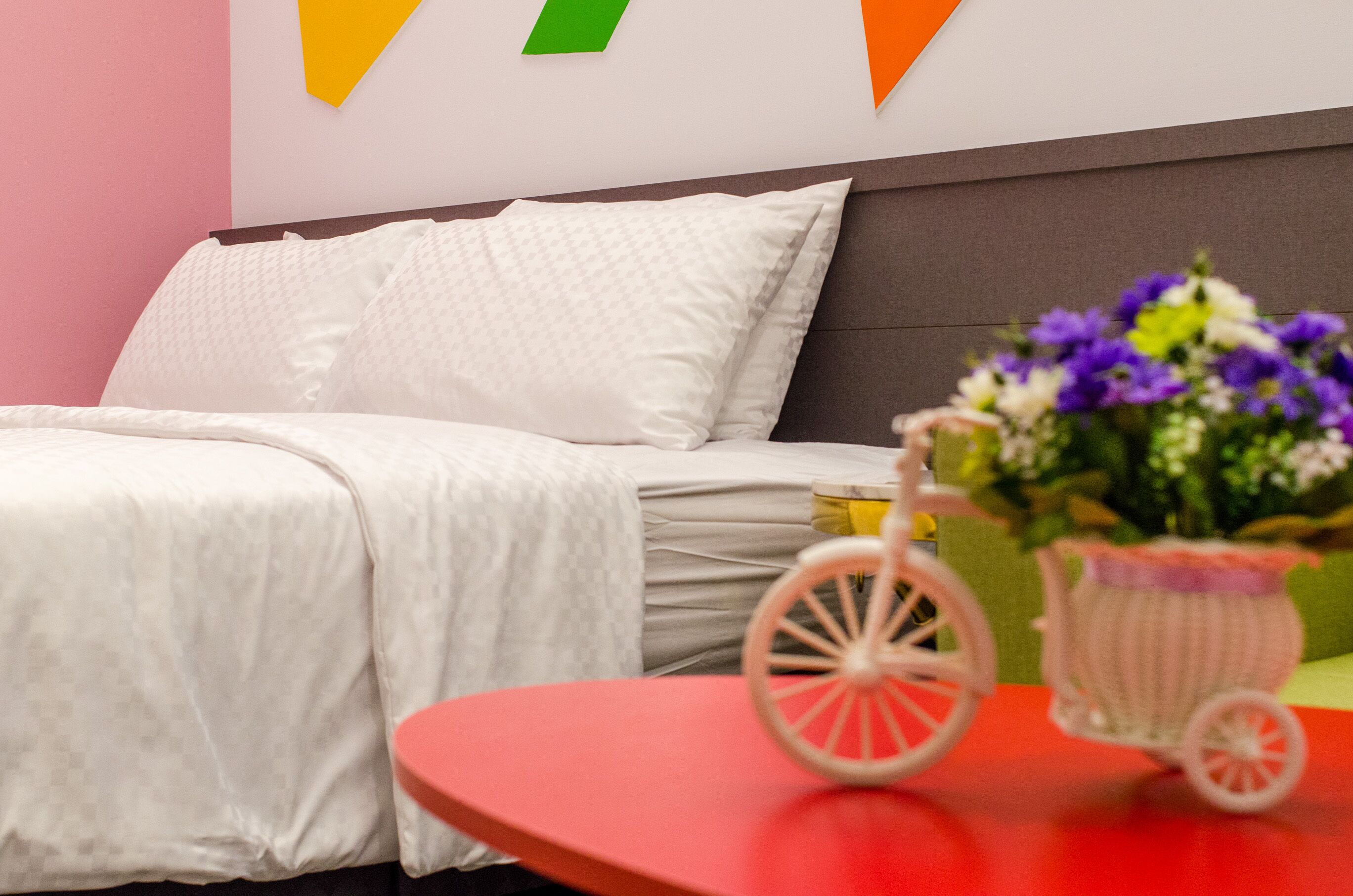 A neatly made bed with white bedding, a small table with a pink toy car and flowers, and a wall with colorful triangular decorations.