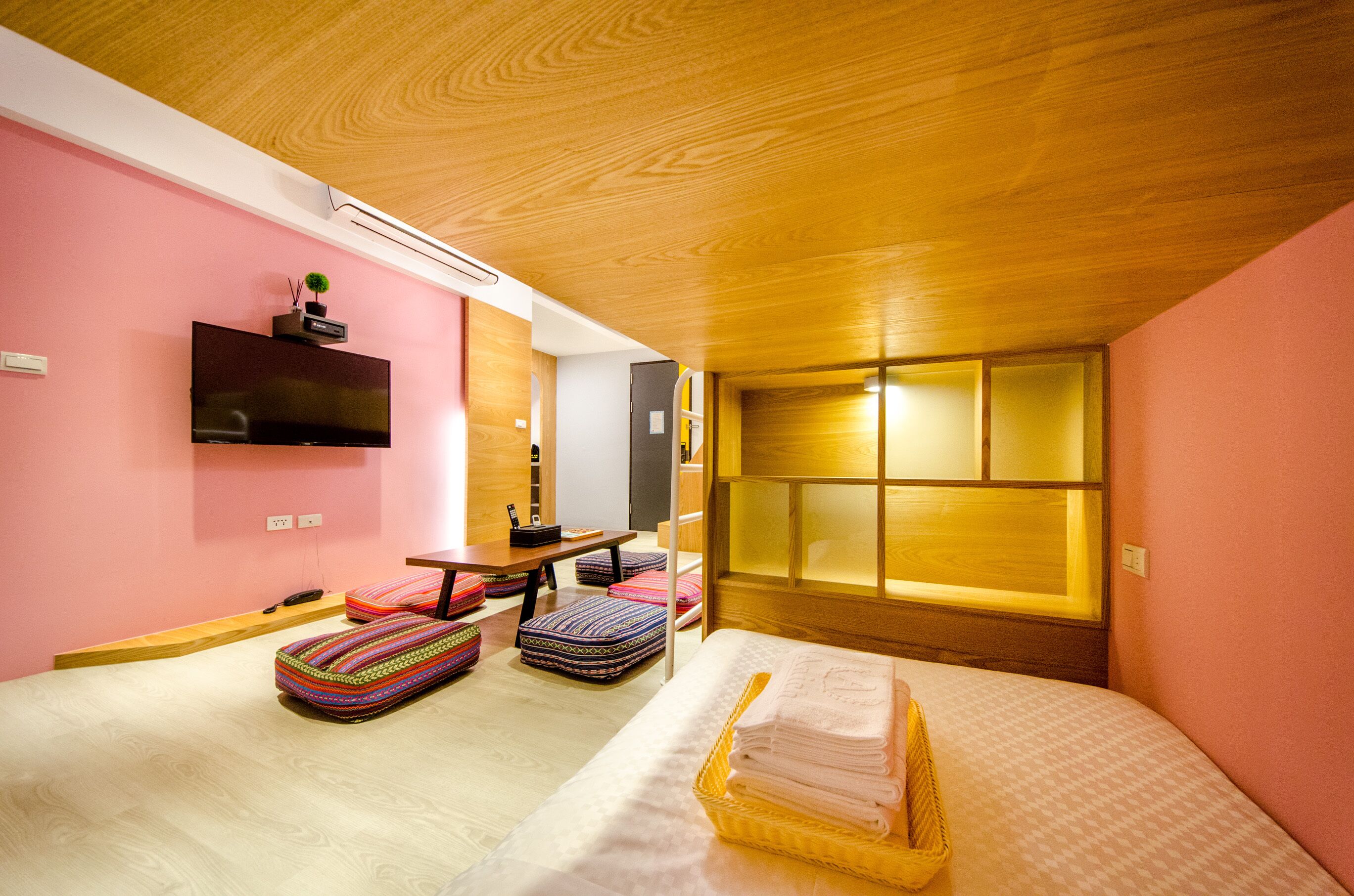 A modern hotel room with a wooden ceiling, pink walls, a flat-screen TV, a bed with a basket of towels, and colorful ottomans.