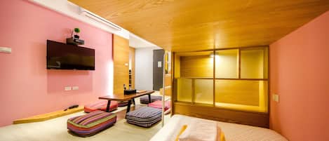 A modern hotel room with a wooden ceiling, pink walls, a flat-screen TV, a bed with a basket of towels, and colorful ottomans.