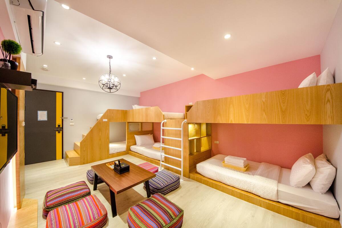 A compact hotel room with bunk beds, a sofa, a small table, and colorful striped ottomans.