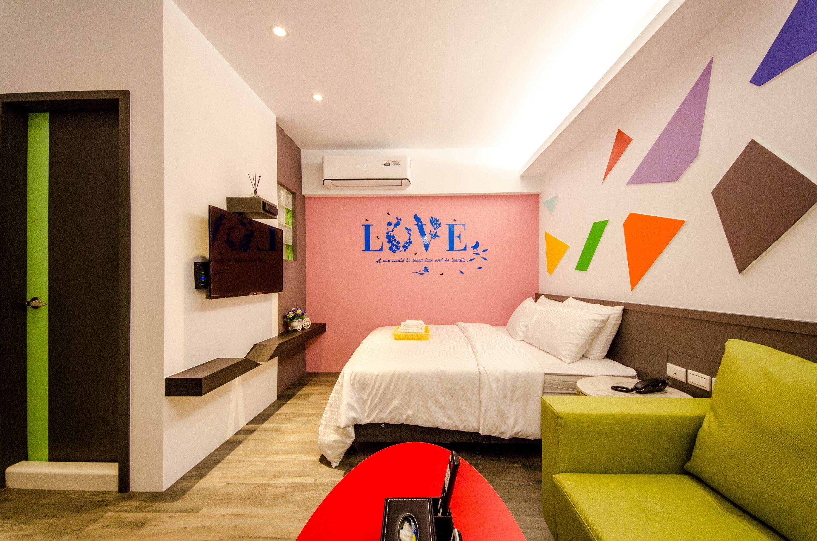 A modern hotel room with a bed, a red chair, a green chair, a wall with geometric shapes, and a 'LOVE' sign.