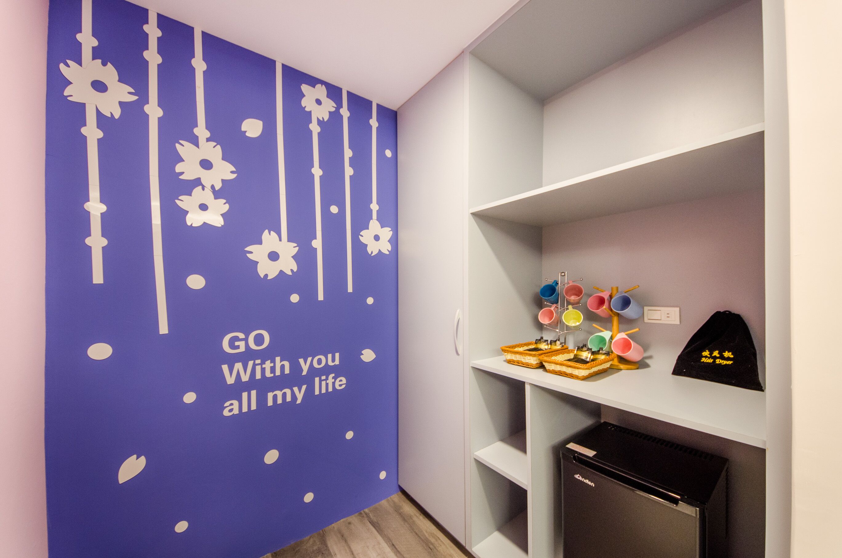 Une pièce avec un mur violet orné d’une fresque représentant l’inscription « Je serai toujours à vos côtés », des étagères blanches et un petit réfrigérateur.