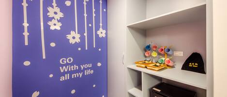 Une pièce avec un mur violet orné d’une fresque représentant l’inscription « Je serai toujours à vos côtés », des étagères blanches et un petit réfrigérateur.