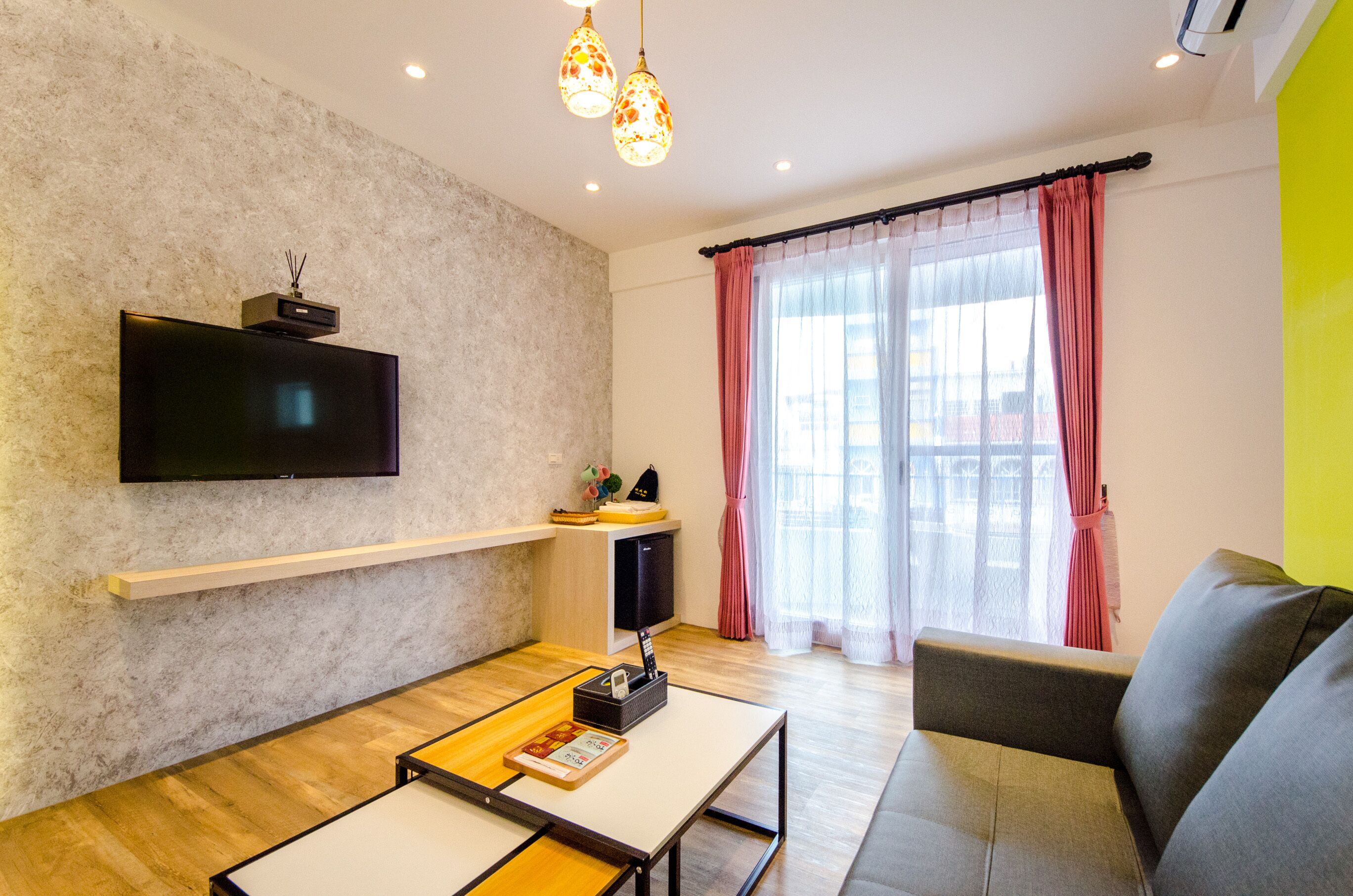 Una sala moderna con una televisión de pantalla plana montada en una pared texturizada, un sofá, una mesa de centro y una ventana con cortinas ligeras.