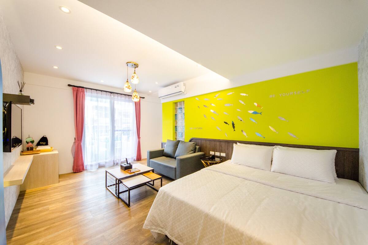 A modern hotel room with a bed, a sofa, a coffee table, and a bright wall with a fish mural.