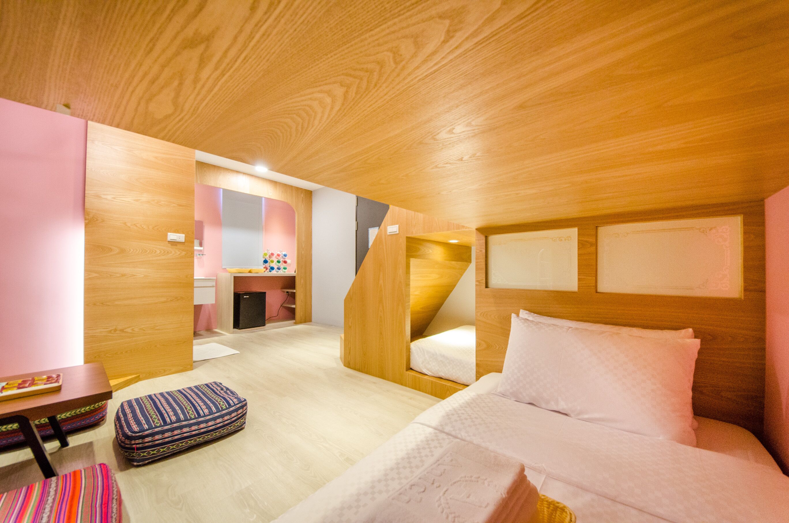 A compact, modern room with a wooden ceiling and walls, a bunk bed, a small desk with a chair, and a colorful rug.