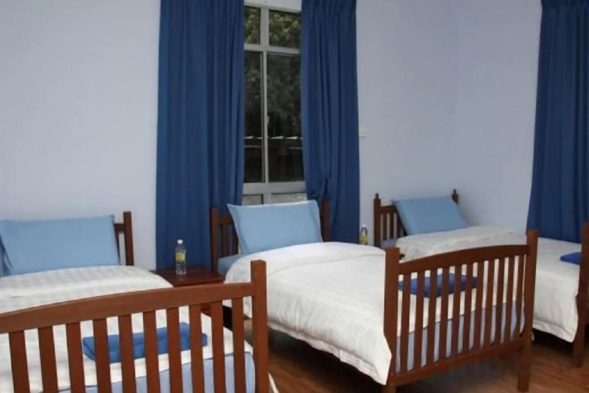 Een kamer met twee houten bedden, elk met een witte matras en blauw beddengoed. Er hangen blauwe gordijnen voor de ramen en op een klein tafeltje staat een fles.