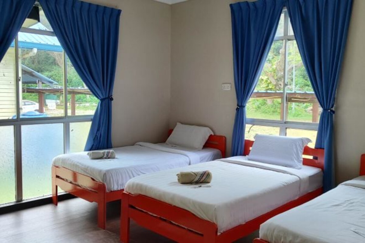 Una habitación con tres camas individuales, cada una con colchón blanco y cabecera roja. La habitación tiene ventanas grandes con cortinas azules, y las paredes están pintadas de un color claro.