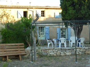 A rustic outdoor seating area with a wooden bench and white plastic chairs under a pergola, overlooking a stone wall and a building with blue shutters.