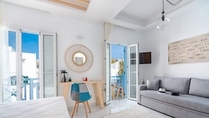 A modern hotel room with a bed, a grey sofa, a desk with a chair, a TV, and a balcony with a view. - Central suites (Mykonos)