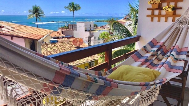 A hammock on a balcony with a view of the ocean and palm trees.