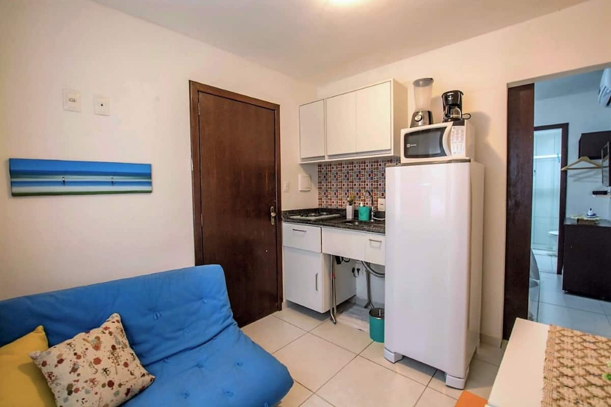 A compact kitchen with white cabinets, a microwave, and a refrigerator. There's a blue sofa with a floral pillow and a small painting on the wall.